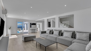 Una sala moderna con un sofá gris, una mesa de centro y un televisor. Hay un ventanal con vista al mar.