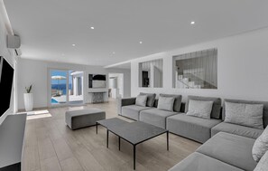 A modern living room with a grey sofa set, a coffee table, and a TV. There is a large window with a view of the sea.