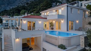 Una casa moderna de dos pisos con piscina, mobiliario exterior y montañas al fondo.