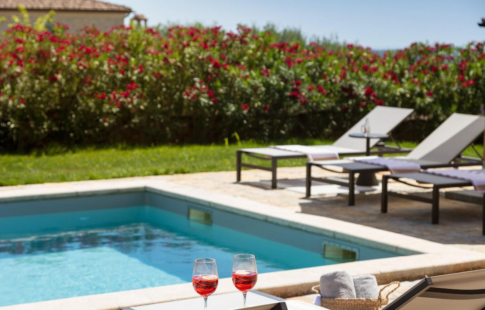 A pool area with lounge chairs and a table, two glasses of red wine, and a basket with towels.