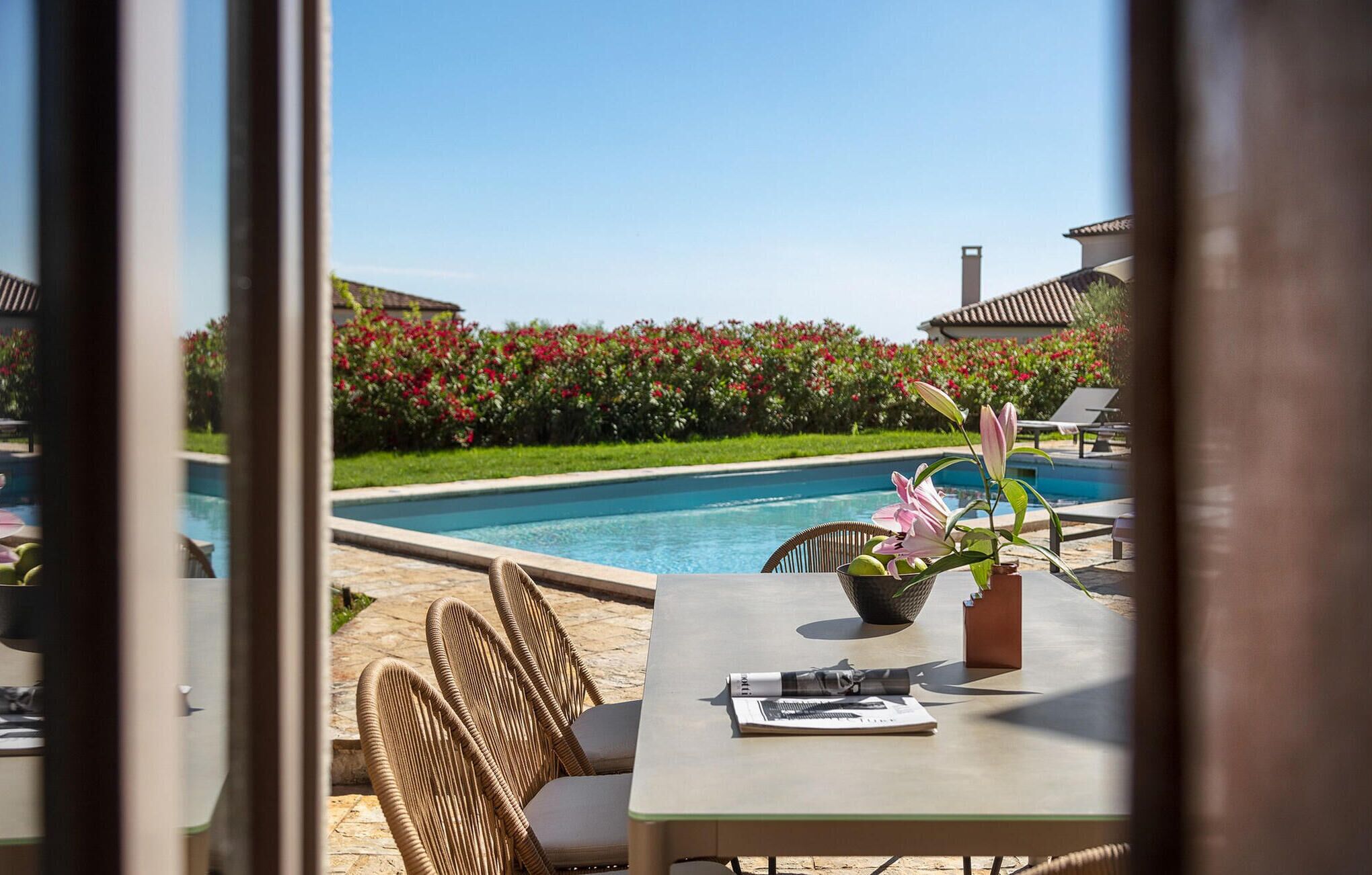 Un espace aménagé au bord de la piscine, avec des fauteuils en osier, une table ornée d’un vase de fleurs, et une vue sur la piscine et une végétation luxuriante.