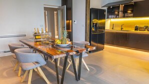 Un espace repas moderne avec une table en bois, quatre chaises grises et un saladier à fruits. On aperçoit à l’arrière-plan une cuisine avec un réfrigérateur noir et un coin bar.