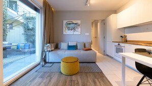 Un salon moderne et bien éclairé, avec un canapé, un pouf jaune et une grande fenêtre donnant sur l’extérieur.