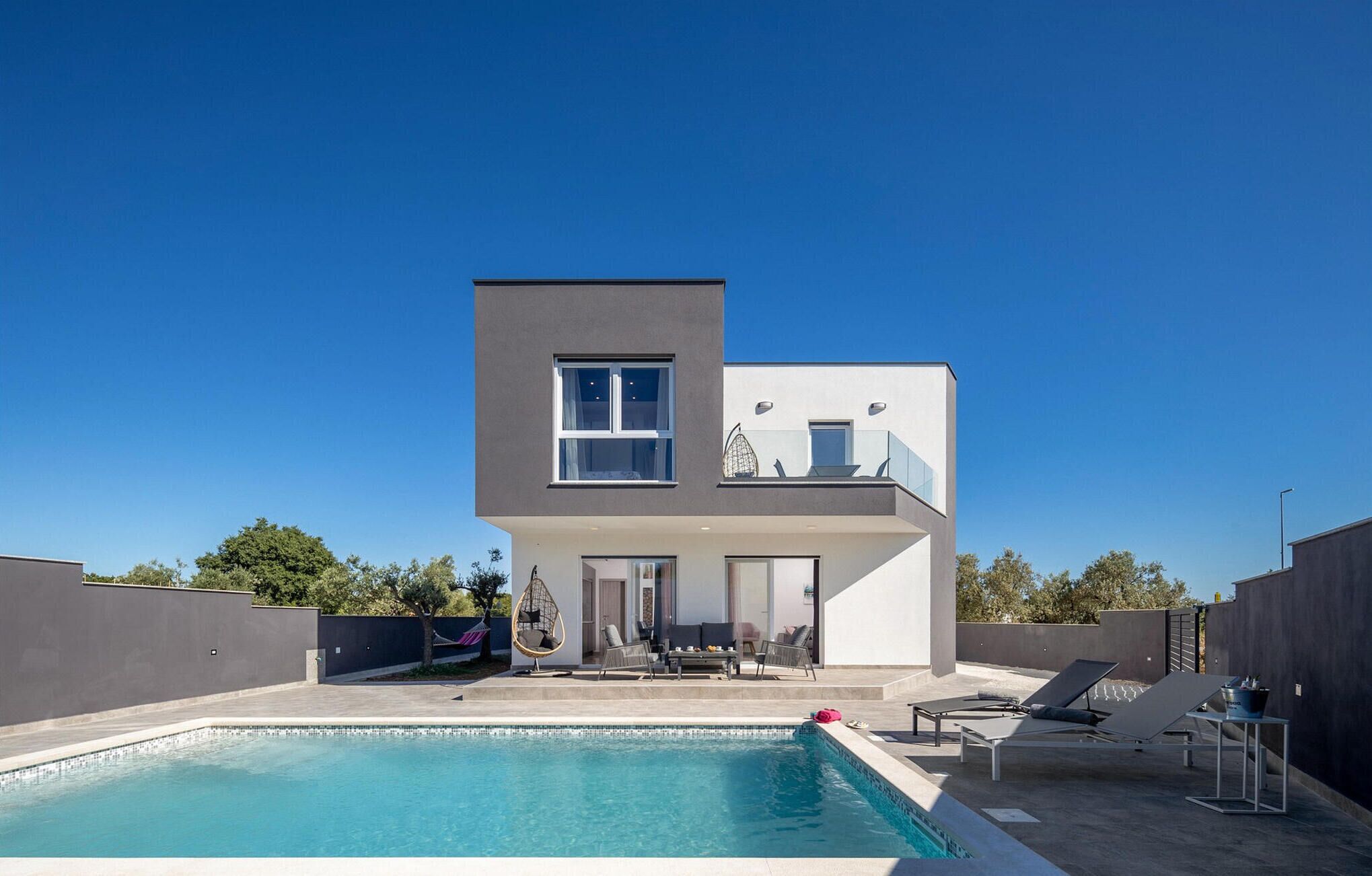 A modern two-story house with a swimming pool, outdoor seating, and a clear blue sky.