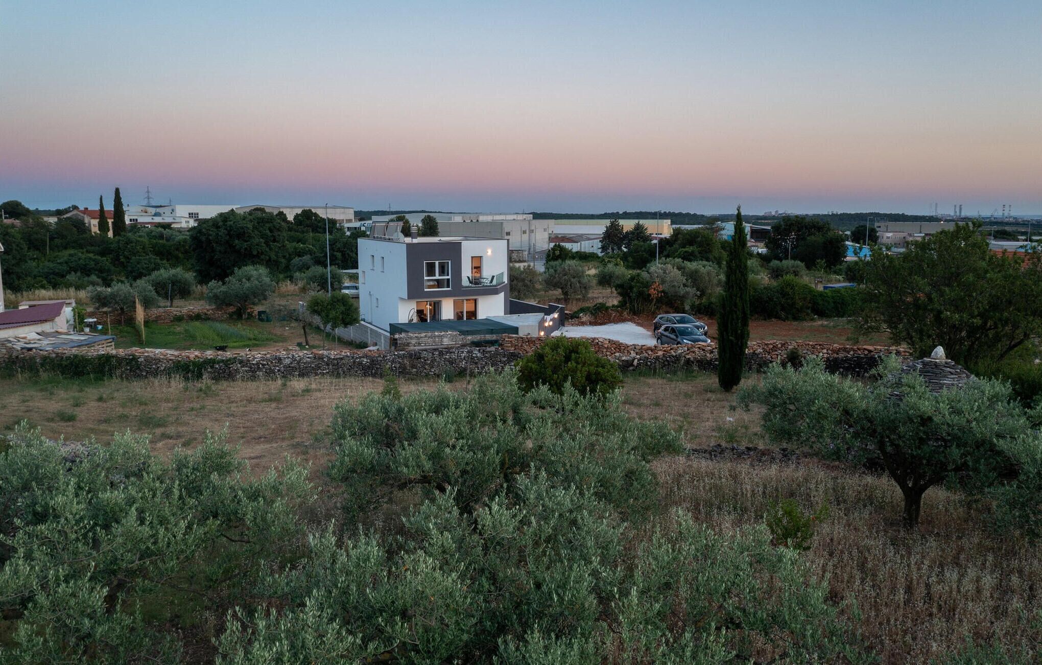 A modern two-story house with a balcony, surrounded by olive trees and a stone wall.