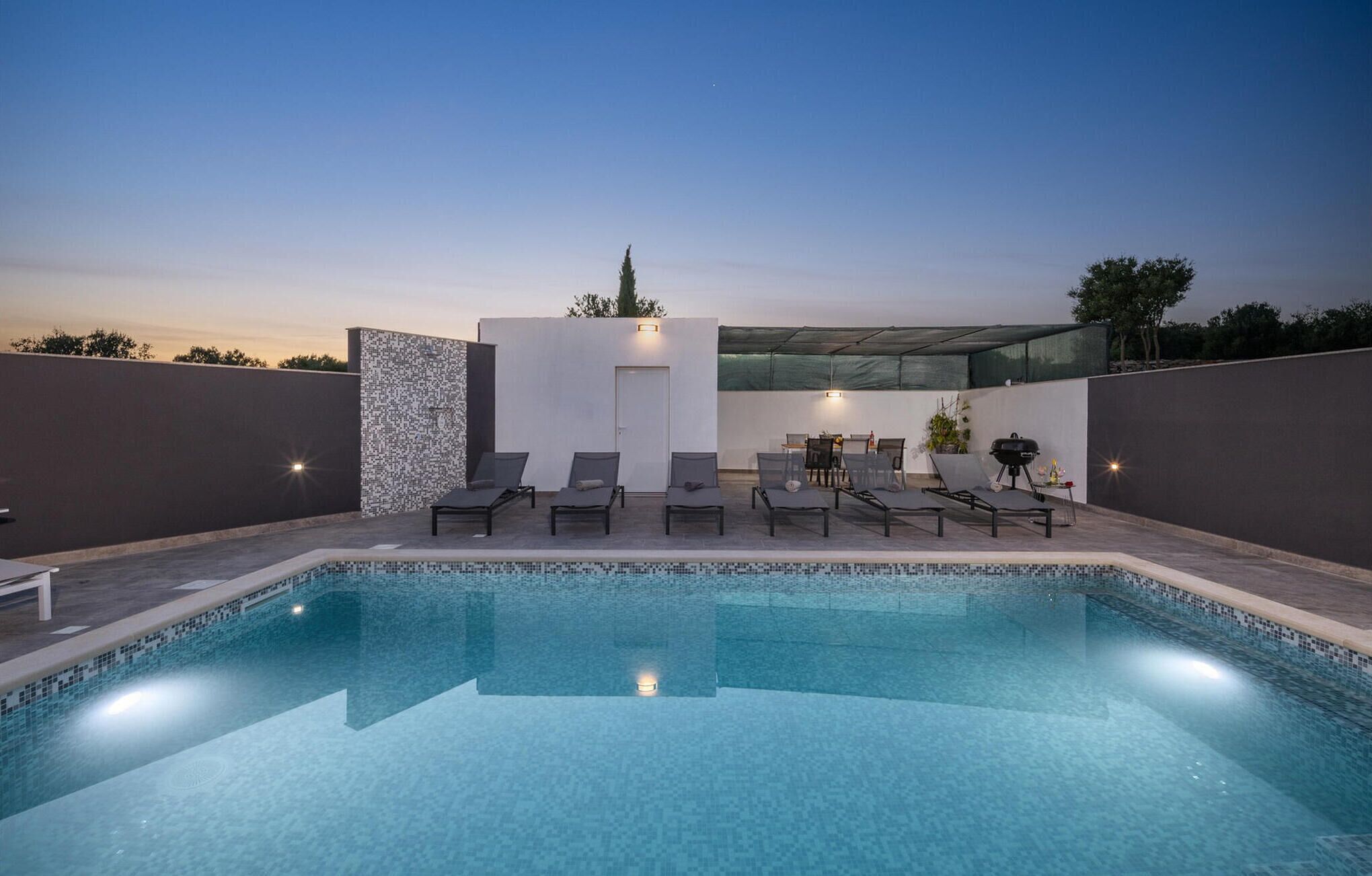 A modern outdoor pool area with lounge chairs and a barbecue setup.