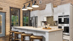 Een moderne keuken met witte kastjes, een groot eiland met krukken, roestvrijstalen apparatuur en een uitzicht op de buitenlucht door grote ramen.
