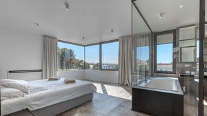 Un dormitorio moderno con una cama grande, un baño con paredes de vidrio y vistas a la ciudad.