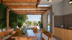 An outdoor patio with wooden furniture, a table, and a bench. There is a view of a swimming pool and greenery in the background.