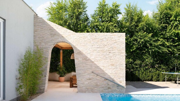 A modern outdoor pool area with a stone wall, a wooden bench, and a potted plant.