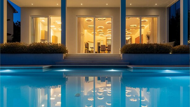 Une maison moderne avec une piscine à l’avant, de grandes portes vitrées et un intérieur bien éclairé.