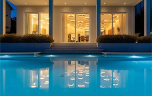 Une maison moderne avec une piscine à l’avant, de grandes portes vitrées et un intérieur bien éclairé.