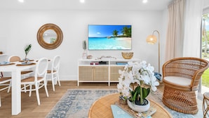 En moderne stue med et hvidt spisebordssæt, en fletstol, et stort fladskærms-tv der viser et strandbillede, og et fletsidebord med dekorative genstande.