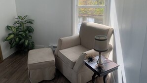 Et hjørneværelse med en beige lænestol, skammel i samme farve, et sengebord med lampe og en stor stueplante.