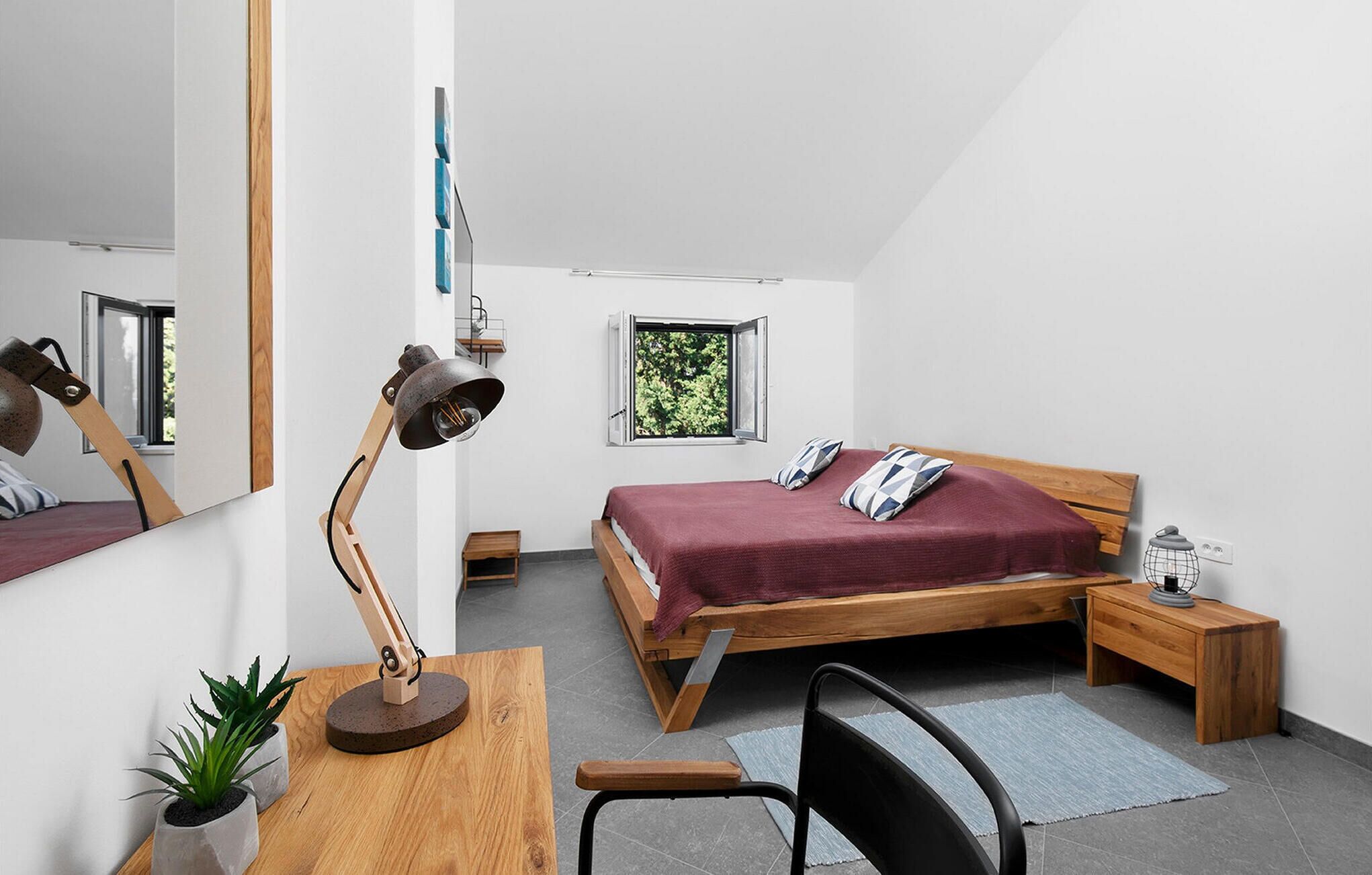 Ein modernes Schlafzimmer mit einem hölzernen Bett, einem Schreibtisch mit Lampe, einem Stuhl, einem kleinen Tisch und einem Fenster mit Blick auf Bäume.