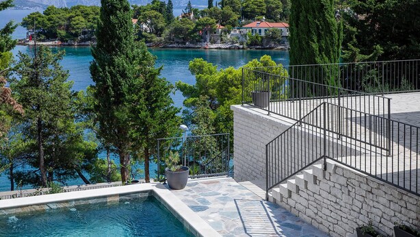 A pool with a stone border, a white brick wall, and a metal railing overlooking a body of water with trees and buildings in the background. - Nice home in Splitska (Splitska)