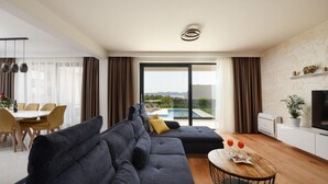 En moderne stue med sofa, spiseområde og udsigt til et pool og bjerge.