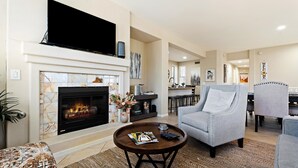 A living room with a fireplace, a television mounted above it, and a dining area in the background. - Peaceful Kierland 2BR Condo | Pool, Spa | Free Parking | Golf Access | Gym Walk to Shops & Dining (Scottsdale)