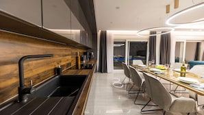 Una cocina moderna con protector de salpicadero de madera, un lavamanos negro y una zona de comedor con mesa y sillas de vidrio.