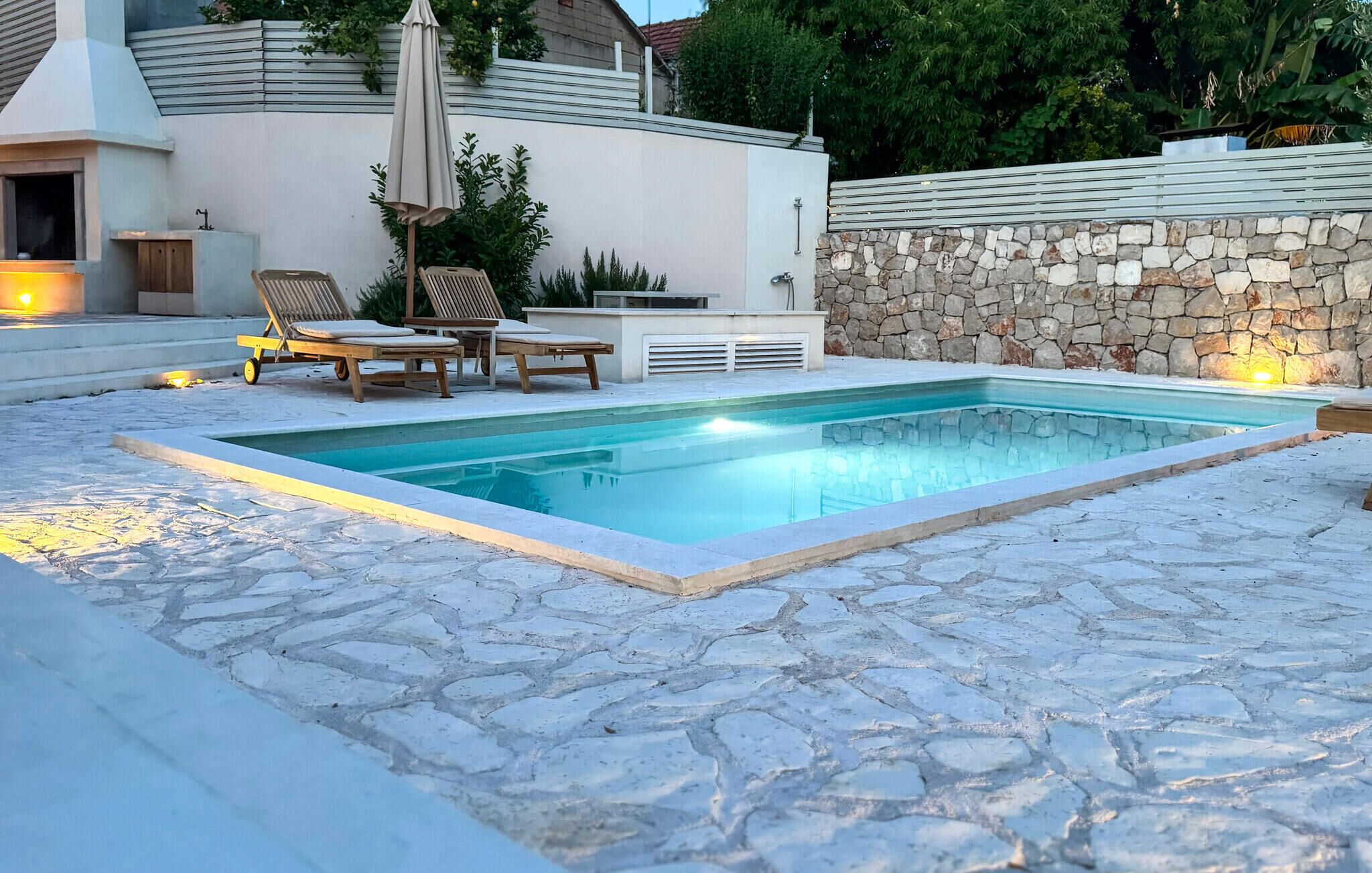 Ein Schwimmbecken mit Steinplatten, Liegestühlen und einer Steinmauer.