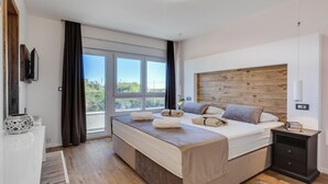 Une chambre moderne avec un grand lit, une tête de lit en bois, une table de chevet et une vue sur l’extérieur grâce à une grande fenêtre.