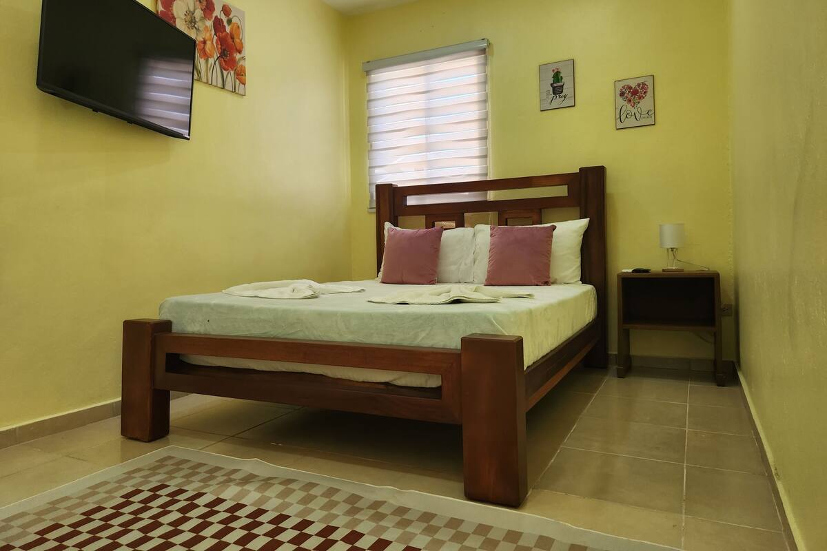 Une chambre avec un lit en bois, une table de chevet avec une lampe, un téléviseur fixé au mur et une fenêtre avec des stores.