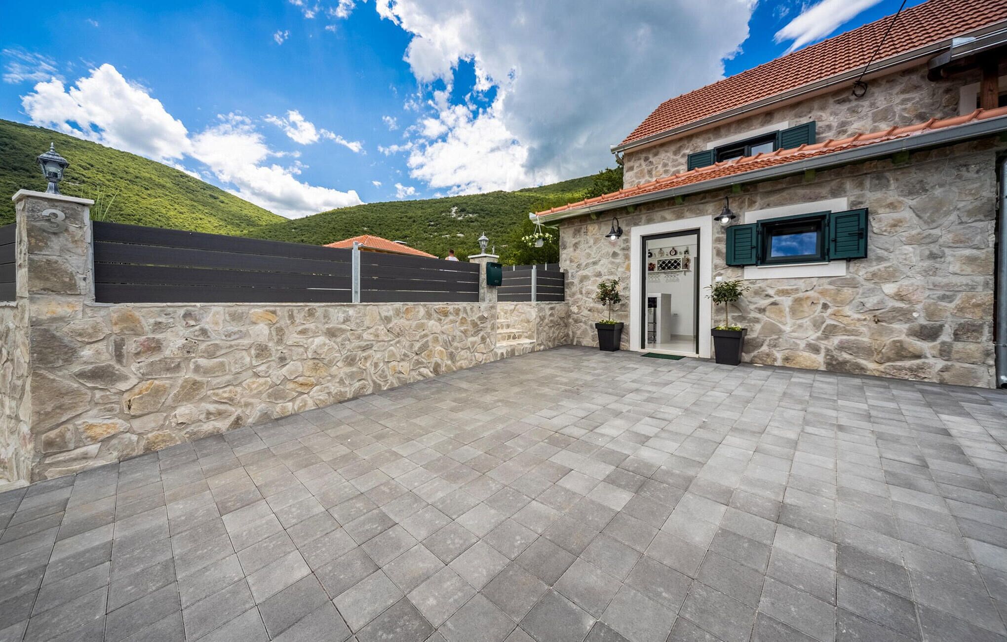 A stone building with a tiled roof, a paved courtyard, and a view of a green hill.