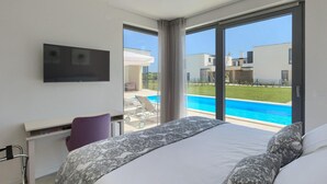 Ein modernes Hotelzimmer mit Bett, Schreibtisch und Fernseher. Es hat eine große Glasschiebetür, die zu einem Swimmingpool und einer Aussicht auf die Umgebung führt.
