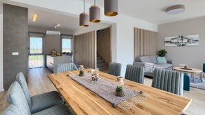 Ein modernes Wohnzimmer mit einem Holzesstisch, grau gepolsterten Stühlen und einem grauen Sofa. Über dem Tisch befinden sich Pendelleuchten und eine Treppe, die in den oberen Stockwerk führt.