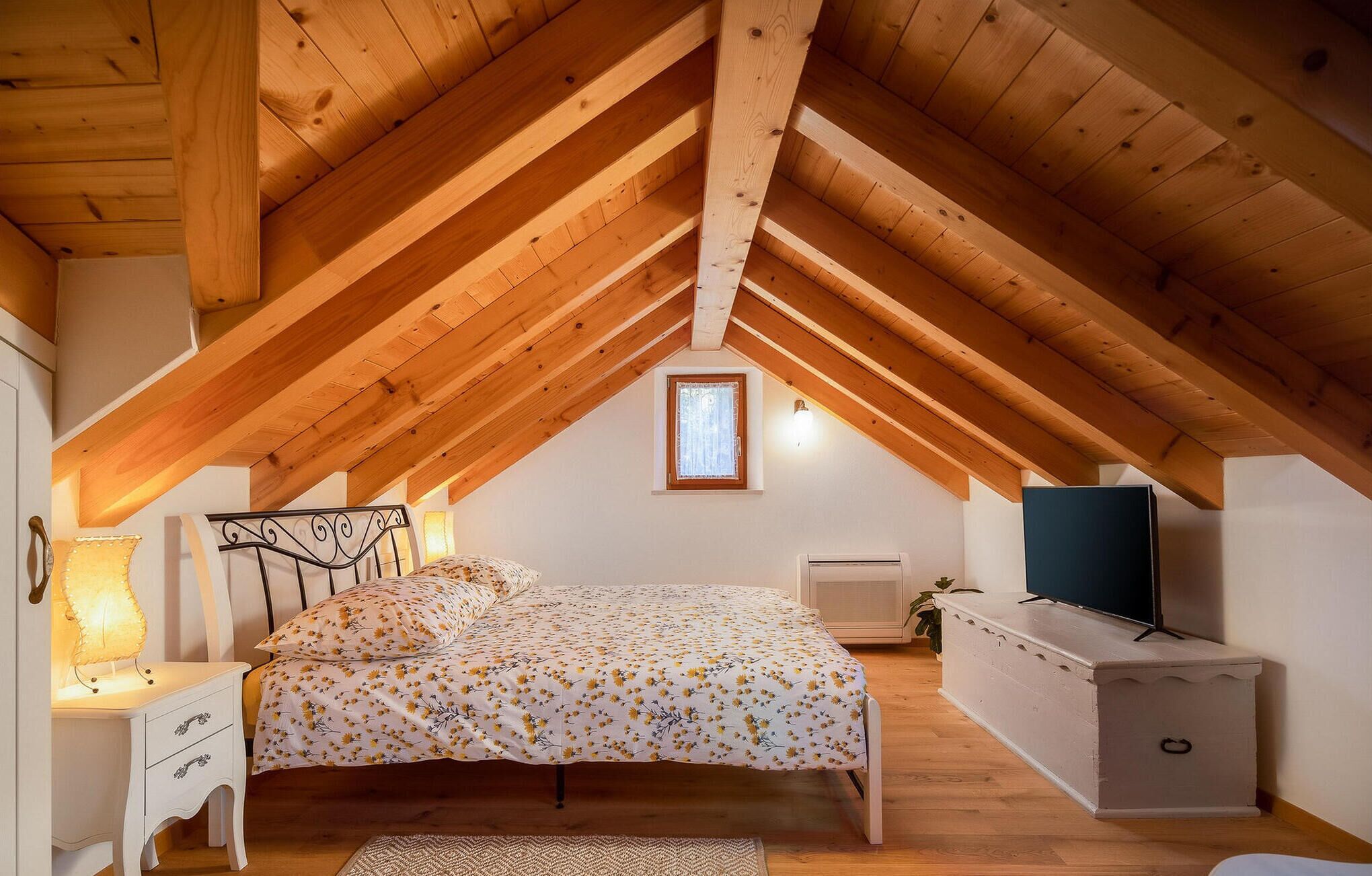 A bedroom with a sloped wooden ceiling, a bed with a floral bedspread, a bedside lamp, a dresser, a television, and a small window.