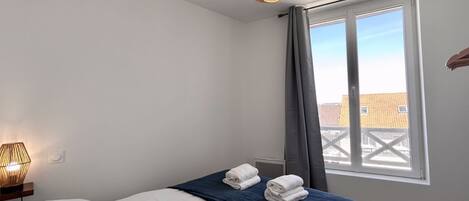 Un dormitorio con cama, lámpara de noche, ventana con cortinas y vistas a edificios.