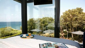 Un lit avec un livre, un plateau avec de la nourriture et une vue sur la mer et les arbres.