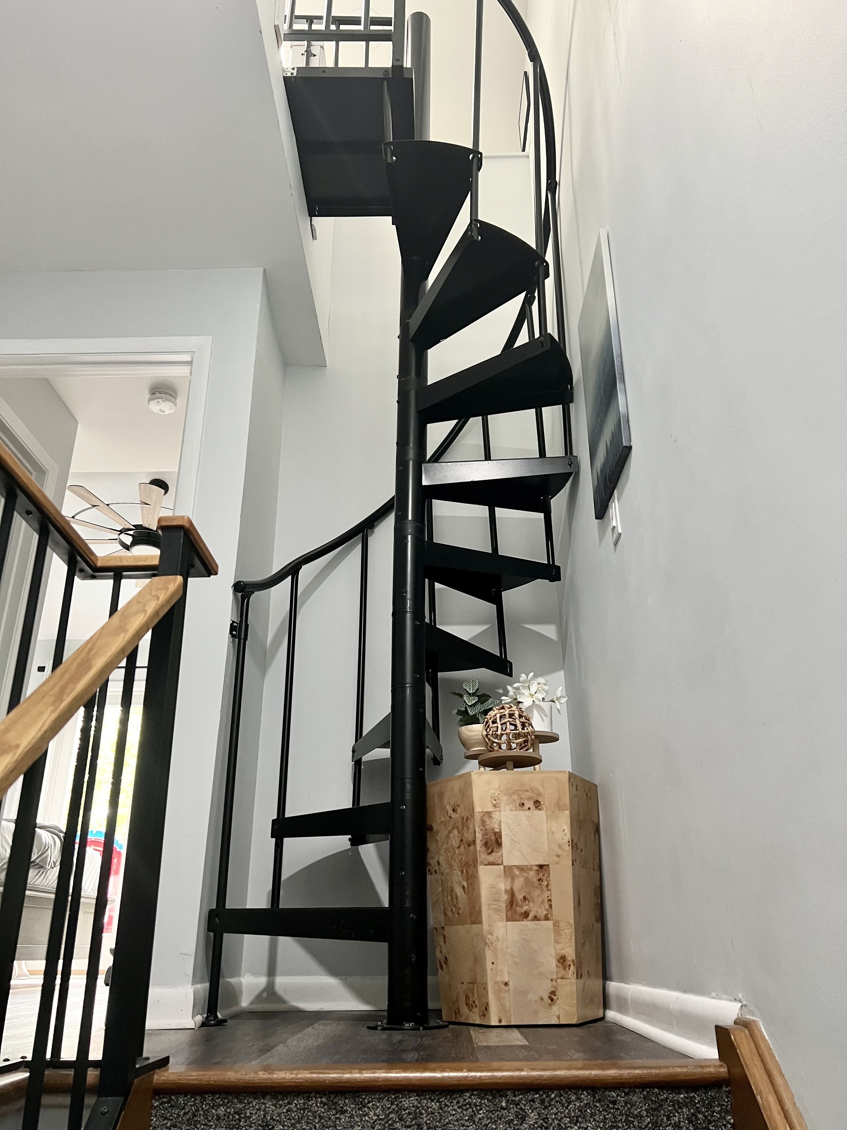 Un escalier en colimaçon noir, doté d’une rampe en bois, et un mur blanc orné d’un tableau.