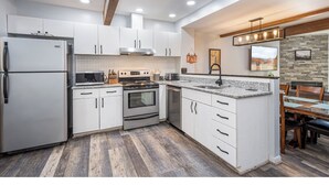 A modern kitchen with white cabinets, stainless steel appliances, a granite countertop, and a dining area with a wooden table and chairs. - 4 Seasons Wisp - Ski In/Ski Out Townhome with Lake Access, Hot Tub  (McHenry)