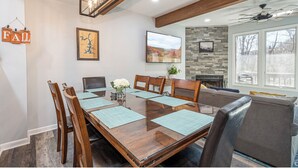 A dining area with a wooden table, chairs, and a glass top. There is a fireplace, a large window, and a ceiling fan. - 4 Seasons Wisp - Ski In/Ski Out Townhome with Lake Access, Hot Tub  (McHenry)