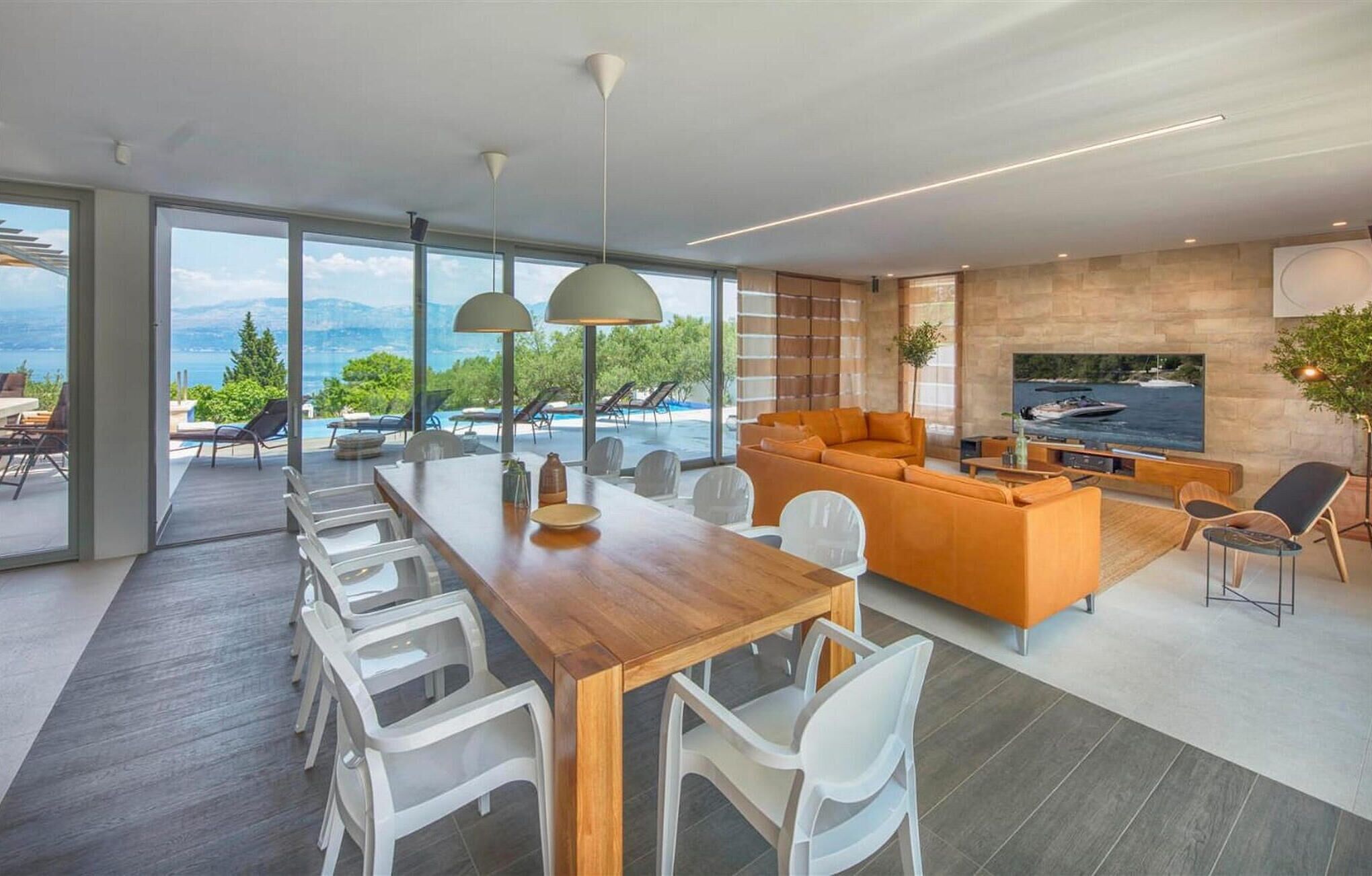 A modern living room with a wooden dining table, white chairs, a large orange sofa, and a TV.