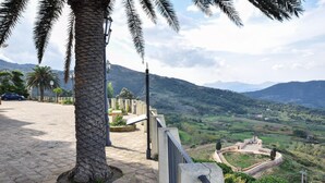 Un sentiero lastricato con una palma e un muro in pietra, affacciato su una valle con una struttura simile a un forte.