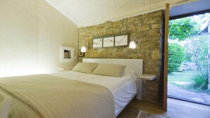 Een slaapkamer met een stenen muur, een bed met wit beddengoed, een nachtkastje en uitzicht op groen.