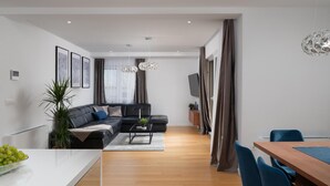 A modern living room with a black sectional sofa, a wooden dining table with blue chairs, a coffee table, and a large window with curtains. - Beautiful home in Privlaka with sauna (Privlaka)