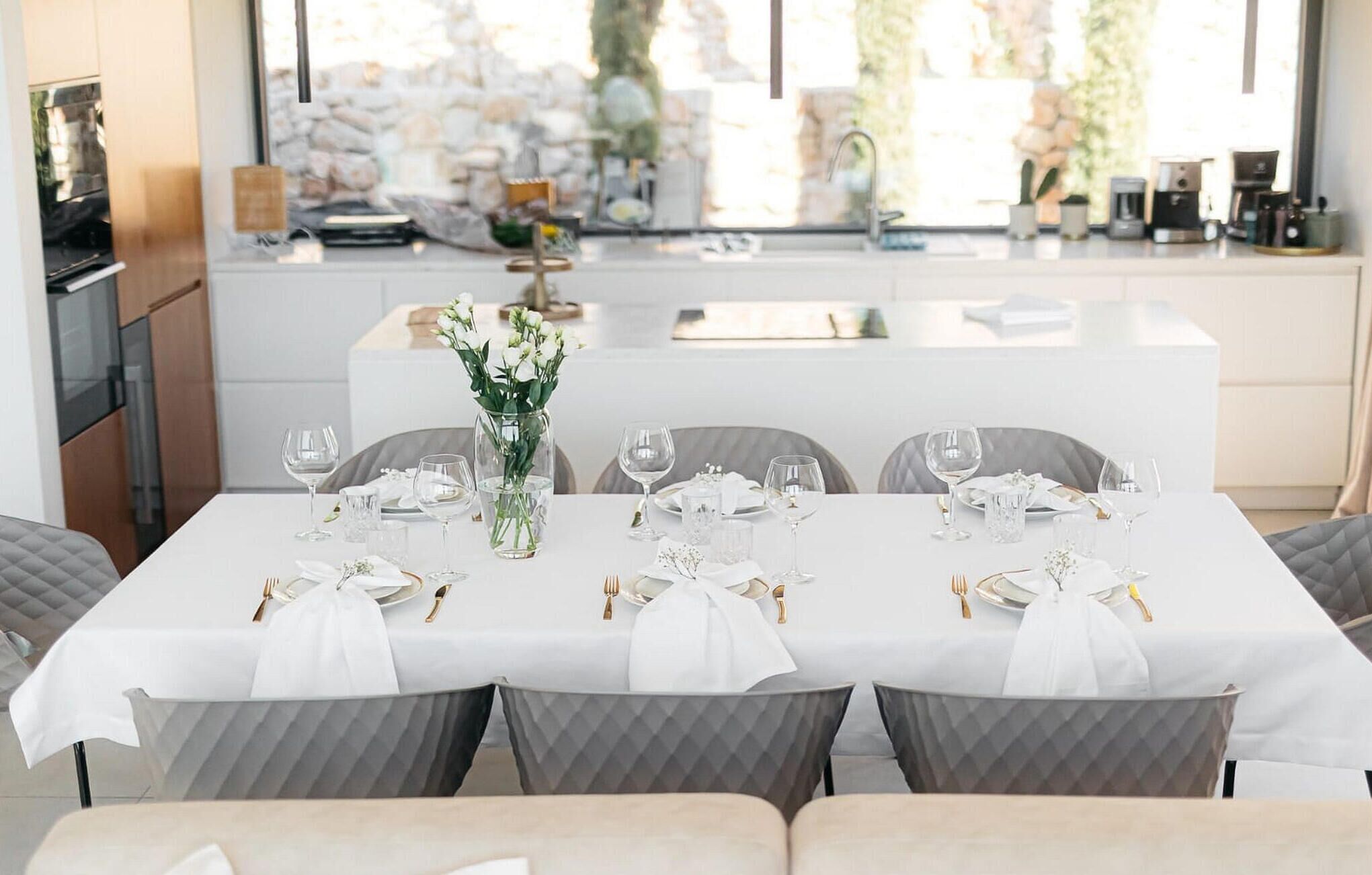 A dining area with a white table set for six, grey chairs, and a vase of white flowers.