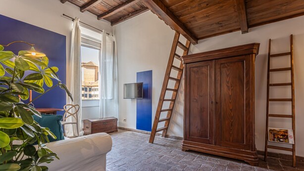Een kamer met een houten plafond, een grote garderobe, een aan de muur bevestigde televisie, een plant en een raam met gordijnen.
