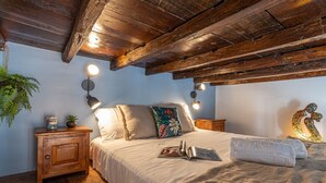Een slaapkamer met een houten plafond, een bed met een witte dekbedovertrek, twee bedlampen, een nachtkastje met een plant en een decoratief wandobject.
