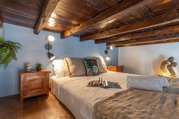A bedroom with a wooden ceiling, a bed with a white bedspread, two bedside lamps, a nightstand with a plant, and a decorative wall piece.