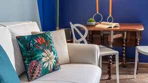 Een woonkamer met een bank, een kleurrijk bloemenkussen, een houten tafel met twee kaarsen en een kleine plant in een pot.