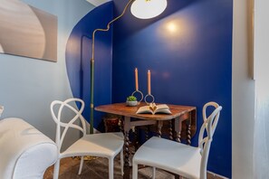 A dining area with a round wooden table, two white chairs, a floor lamp, and two lit candles on a blue wall.