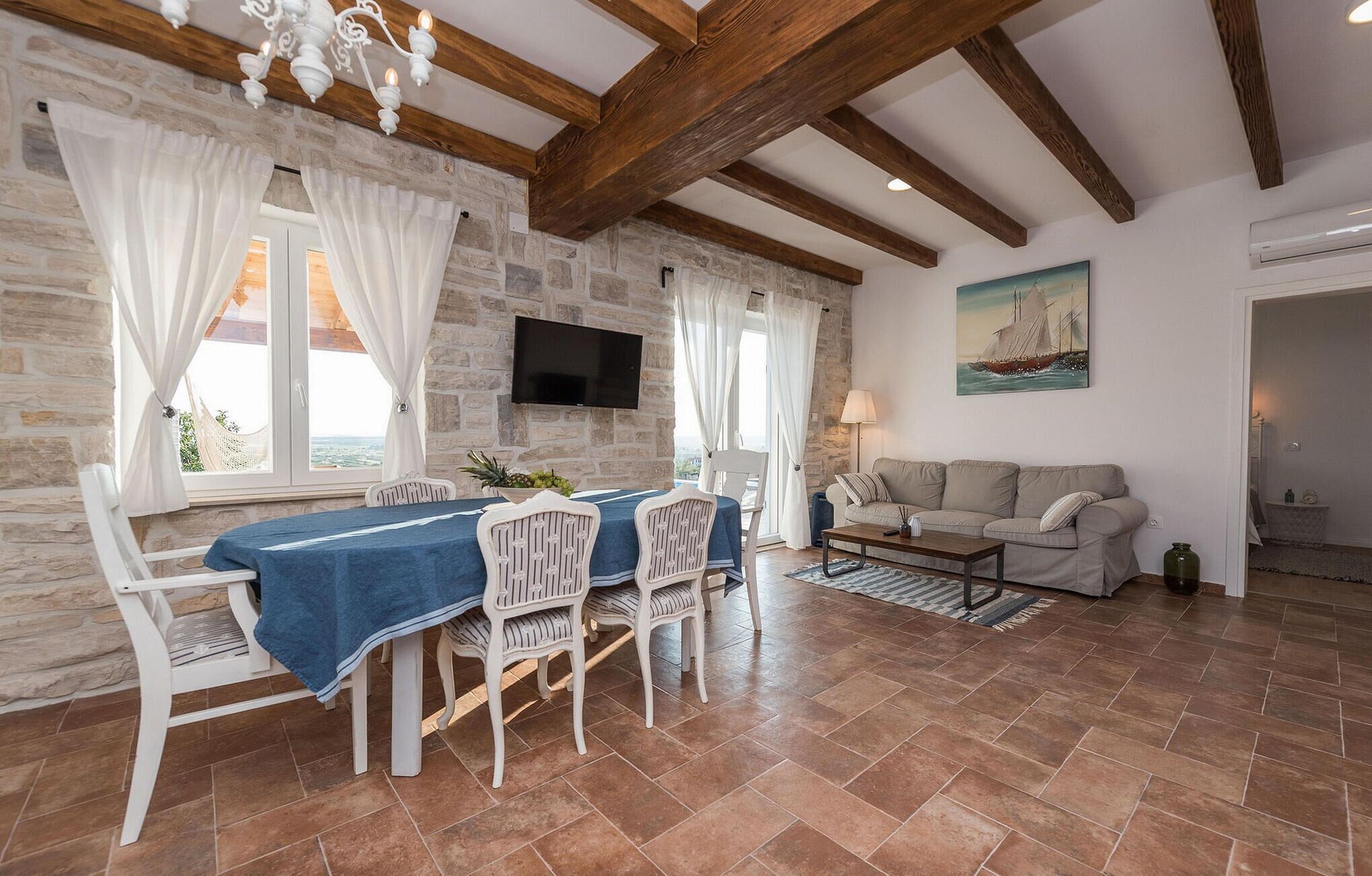Un coin repas avec un mur en pierre, des poutres en bois, un téléviseur fixé au mur, une table avec des chaises et un canapé dans l’espace salon.