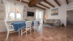 A dining area with a stone wall, wooden beams, a TV mounted on the wall, a table with chairs, and a sofa in a living area. - Villa Mariva (Carro d'oro) (Vrana)