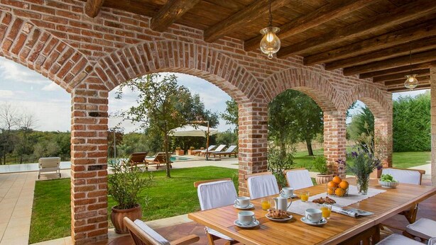 Een terras met buitenmeubilair, voorzien van bakstenen bogen, een houten ontbijttafel en uitzicht op een zwembad en tuin.