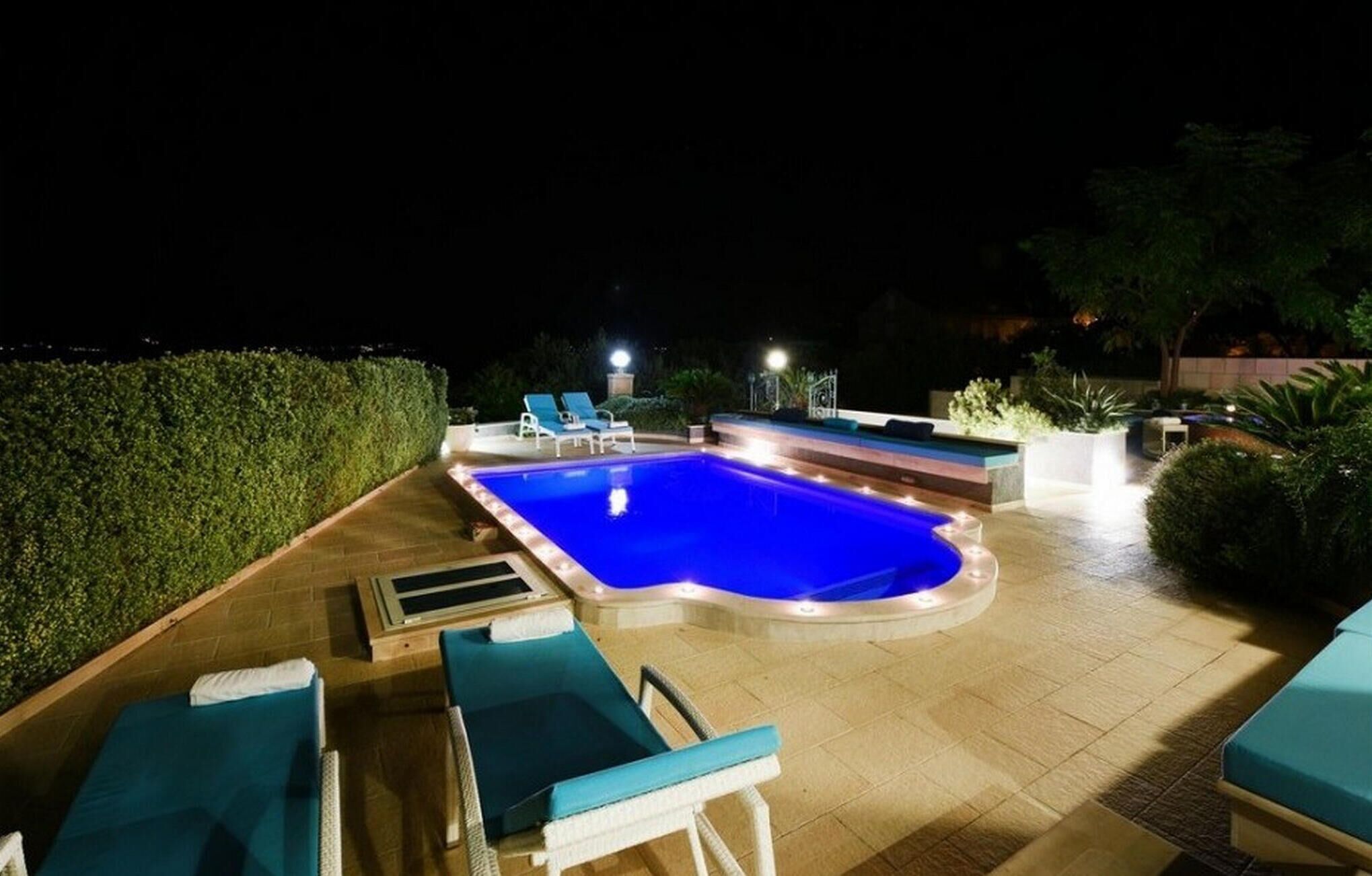 Un área de piscina iluminada durante la noche, con sillones de descanso y una bañera de hidromasaje.
