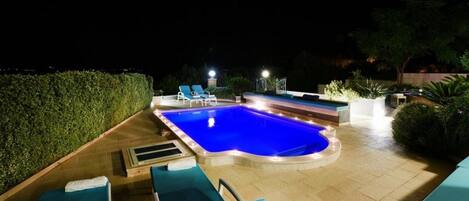 A nighttime swimming pool area with lounge chairs and a hot tub.